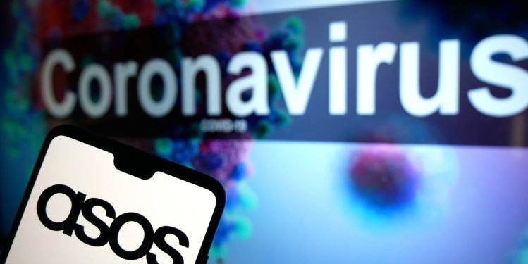 The ASOS logo seen displayed on a mobile phone with an illustrative model of the Coronavirus displayed on a monitor in the background. Photo credit should read: James Warwick/EMPICS Entertainment