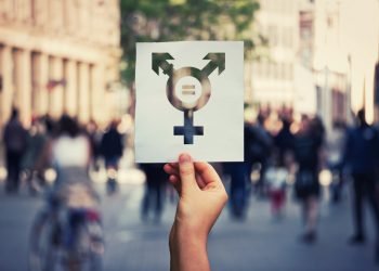 Hand holding a paper sheet with transgender symbol and equal sign inside. Equality between genders concept over a crowded city street background. Sex rights as a metaphor of social issue.