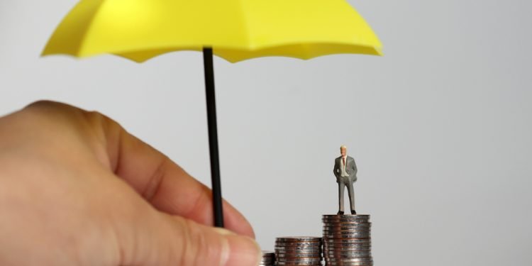 A hand holding a yellow umbrella and a miniature man standing on a pile of coins.