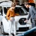 Sales manager showing how to charge electric car to a young client, plugging wire into the car socket at the car dealership