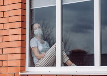 Caucasian woman sitting at window in a medical mask, looking out, wants to go out. protection against coronavirus infection, pandemics, disease outbreaks and epidemics