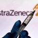 In this photo illustration the medical syringe is seen with AstraZeneca company logo displayed on a screen in the background