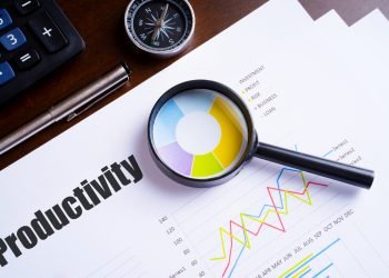 Magnifying glass on colourful pie chart with "Productivity" text on paper, dice, spectacles, pen, laptop calculator on wooden table