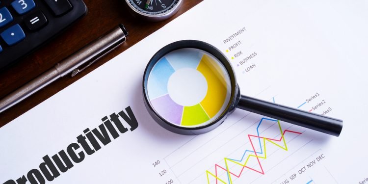 Magnifying glass on colourful pie chart with "Productivity" text on paper, dice, spectacles, pen, laptop calculator on wooden table