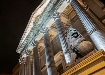 Night view of the main facade of the Palacio de la Cortes, palace of courts, seat of the Congress of Deputies in Madrid, Spain, with one of its iconic bronze lions in the foreground