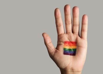 closeup of a rainbow flag in the palm of the hand of a young caucasian person, against an off-white background with some blank space on the left