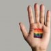 closeup of a rainbow flag in the palm of the hand of a young caucasian person, against an off-white background with some blank space on the left