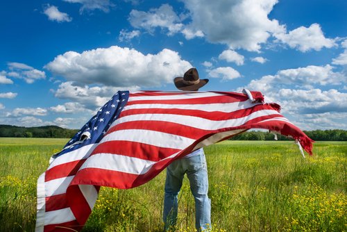 Man wearing cowboy hat waving American flag standing in grass farm agricultural field blue sky white clouds, holidays, patriotism, pride, freedom, conservative, political parties, immigrant, voters