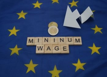 Some coins, next to a white arrow and a wooden inscription, minimum wage, above a flag of the European Union.