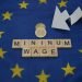 Some coins, next to a white arrow and a wooden inscription, minimum wage, above a flag of the European Union.