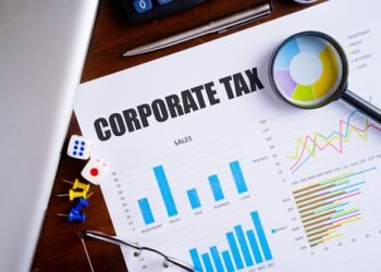 "Corporate Tax" text on paper sheet with magnifying glass on chart, dice, spectacles, pen, laptop and blue and yellow push pin on wooden table - business, banking, finance and investment concept