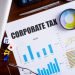 "Corporate Tax" text on paper sheet with magnifying glass on chart, dice, spectacles, pen, laptop and blue and yellow push pin on wooden table - business, banking, finance and investment concept