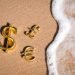 Golden three-dimensional symbols of international dollar, euro and pound currency with wave encroaching on the sand of an empty sunny beach