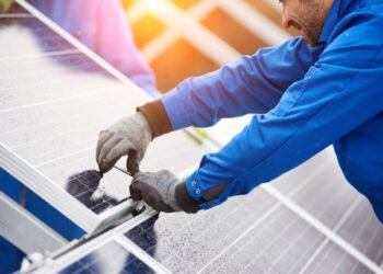 Smiling male technician in blue suit installing photovoltaic blue solar modules with screw. Man electrician panel sun sustainable resources renewable energy source alternative innovation