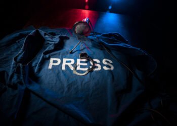 Freedom of the press and journalism concept. Blue journalist (press) vest in dark with backlight and fog. Microphone and handcuffs. Selective focus