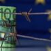 European Union currency wrapped in barbed wire against flag of EU as symbol of Economic warfare, sanctions and embargo busting. Horizontal image. Copy space.