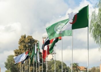 Flags of Algeria, the Arab League and Arab countries flag waving in the wind outside Algiers city with flagposts under a blue cloudy sky and trees in a sunny day.